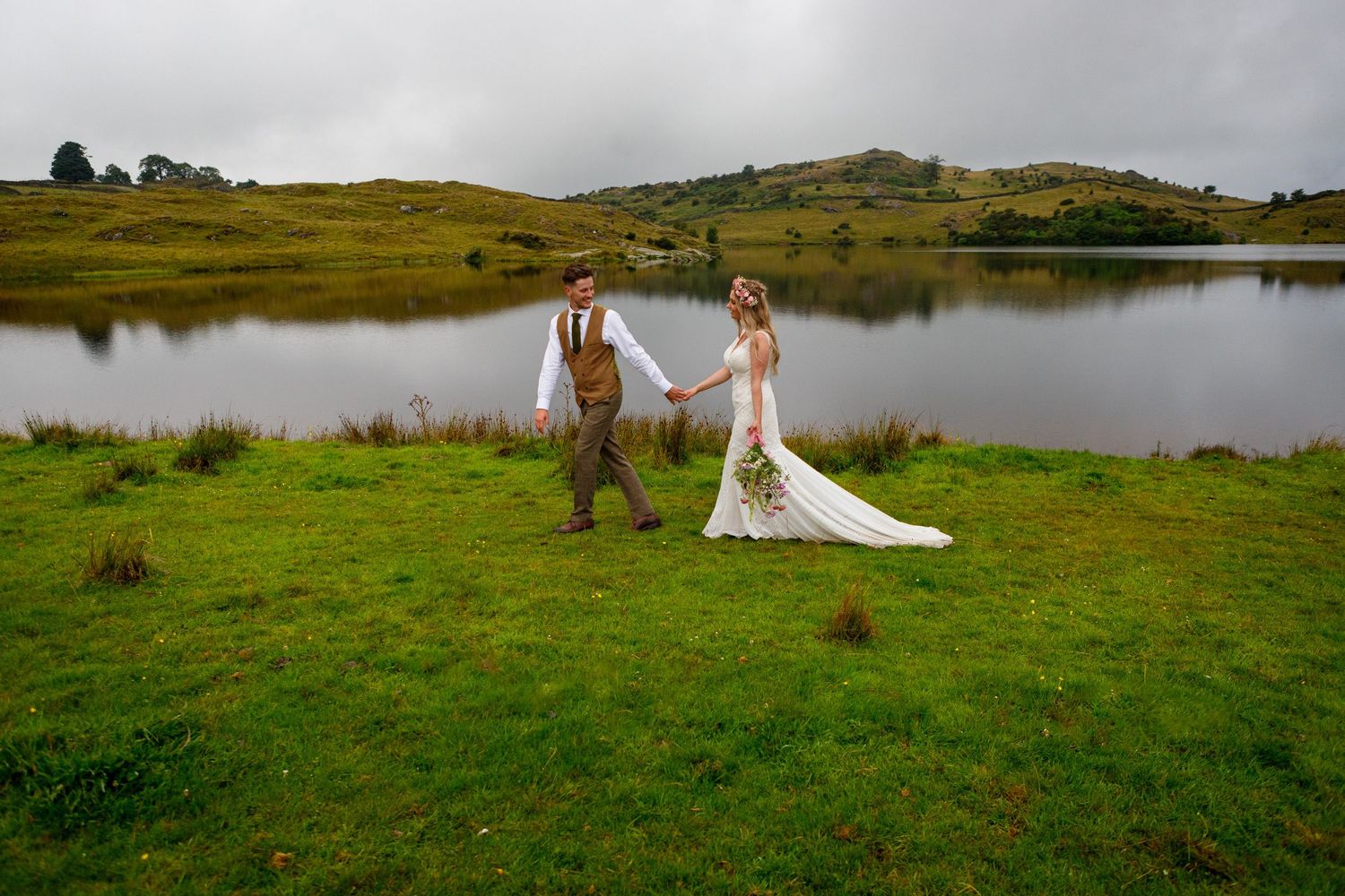 Wedding couple walking by serene lake with mountains and cloudy sky in background.