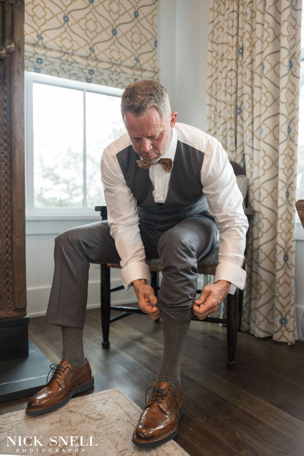 Person in formal attire sitting in a chair while putting on brown leather dress shoes.