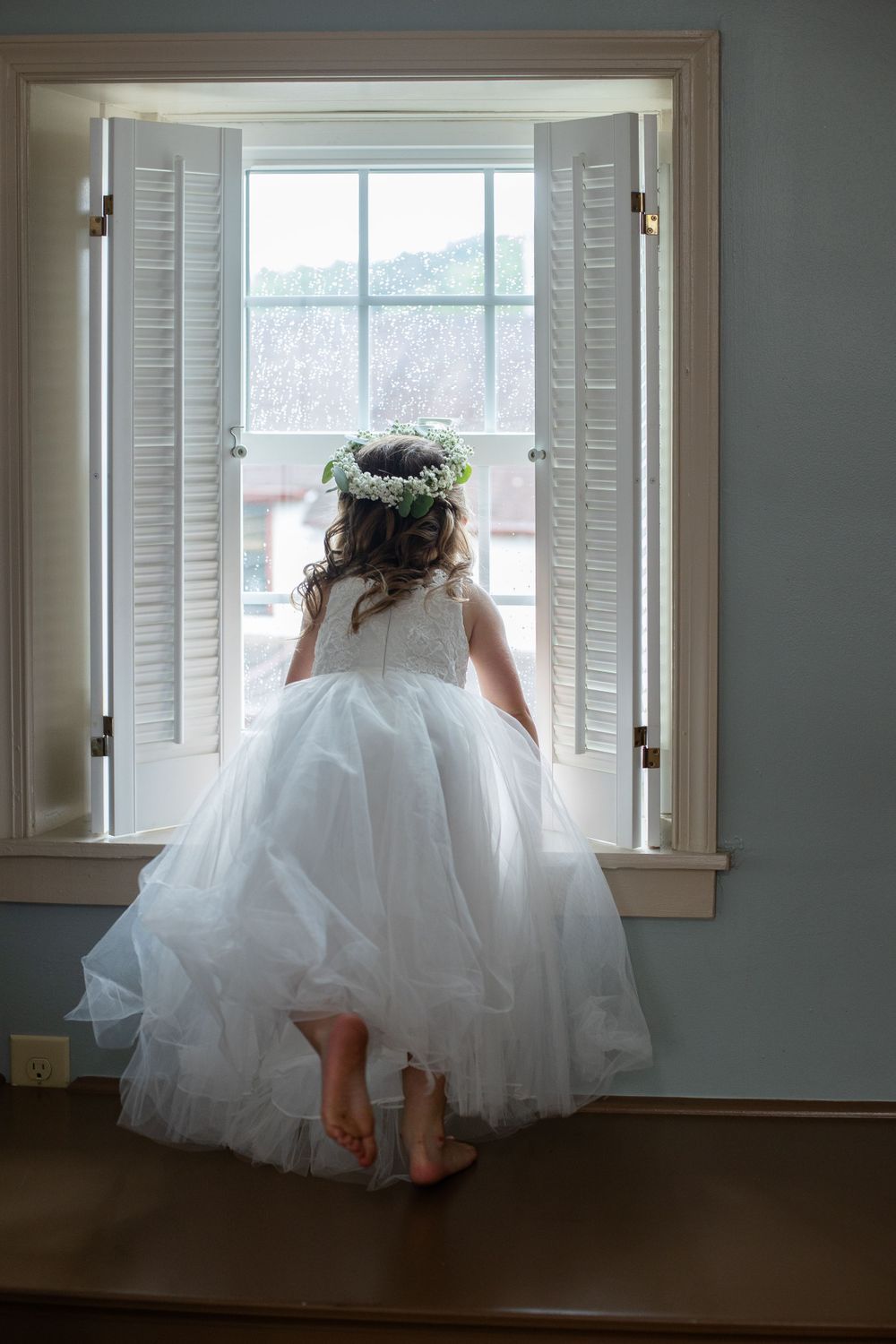 A white flower crown adorns a flowing tulle dress as a figure gazes through plantation shutters by a window.