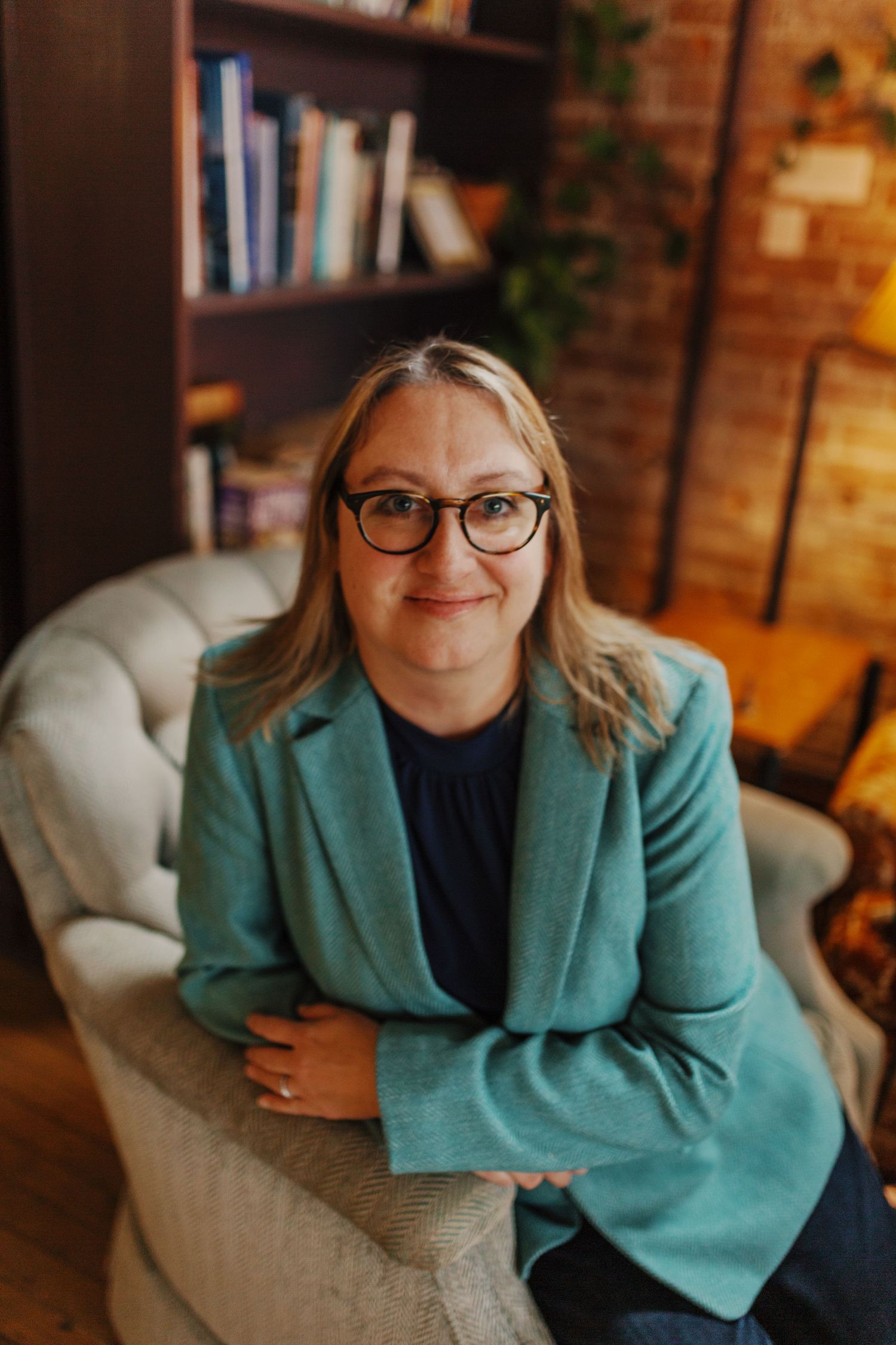 A person wearing glasses and turquoise blazer sits in armchair near bookshelf in cozy home office.