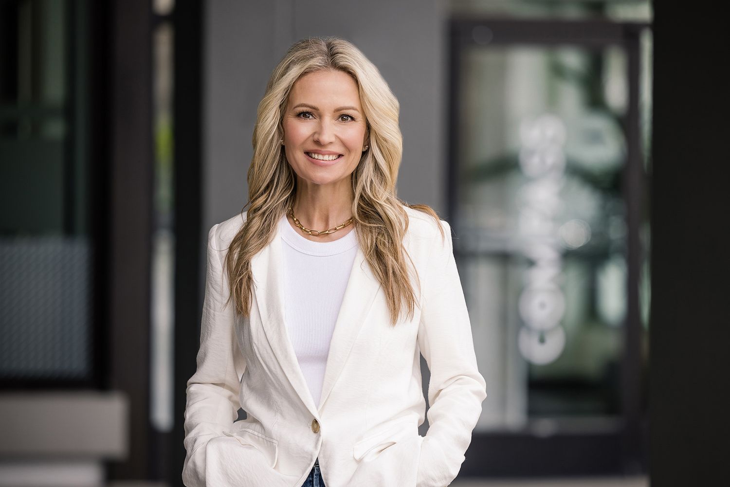 Blonde wavy haired woman wearing white top and white blazer smiling at the camera