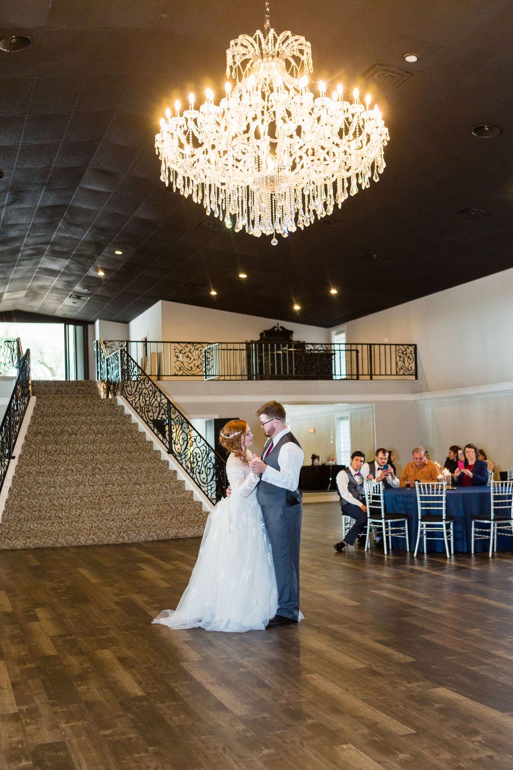 A couple shares their first dance under a crystal chandelier in an elegant ballroom setting.