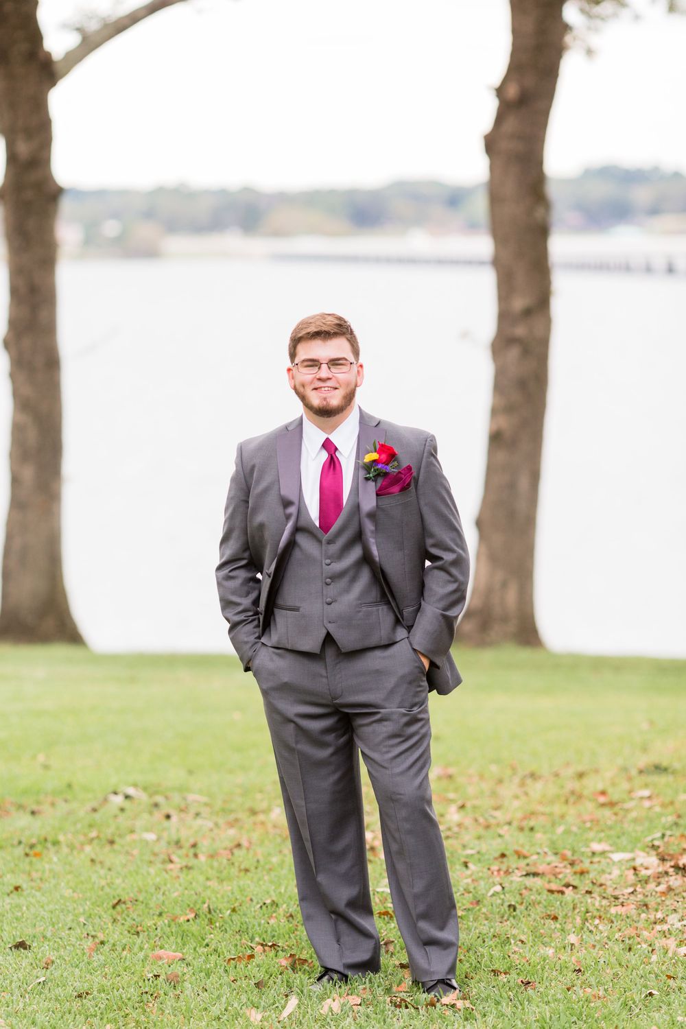Stylish groom in grey suit and pink tie poses by lakefront with trees in background.