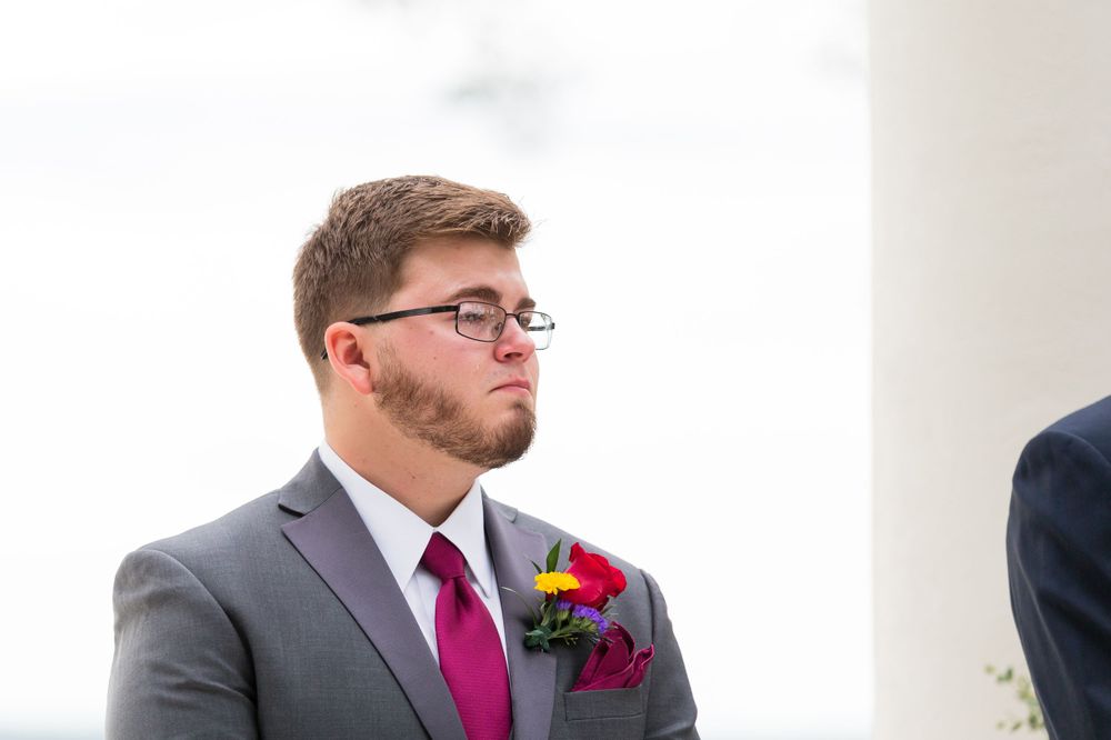 Groom in grey suit with burgundy tie and red boutonniere during outdoor ceremony.