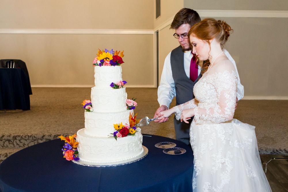 A couple cuts into their white wedding cake decorated with colorful fresh flowers.