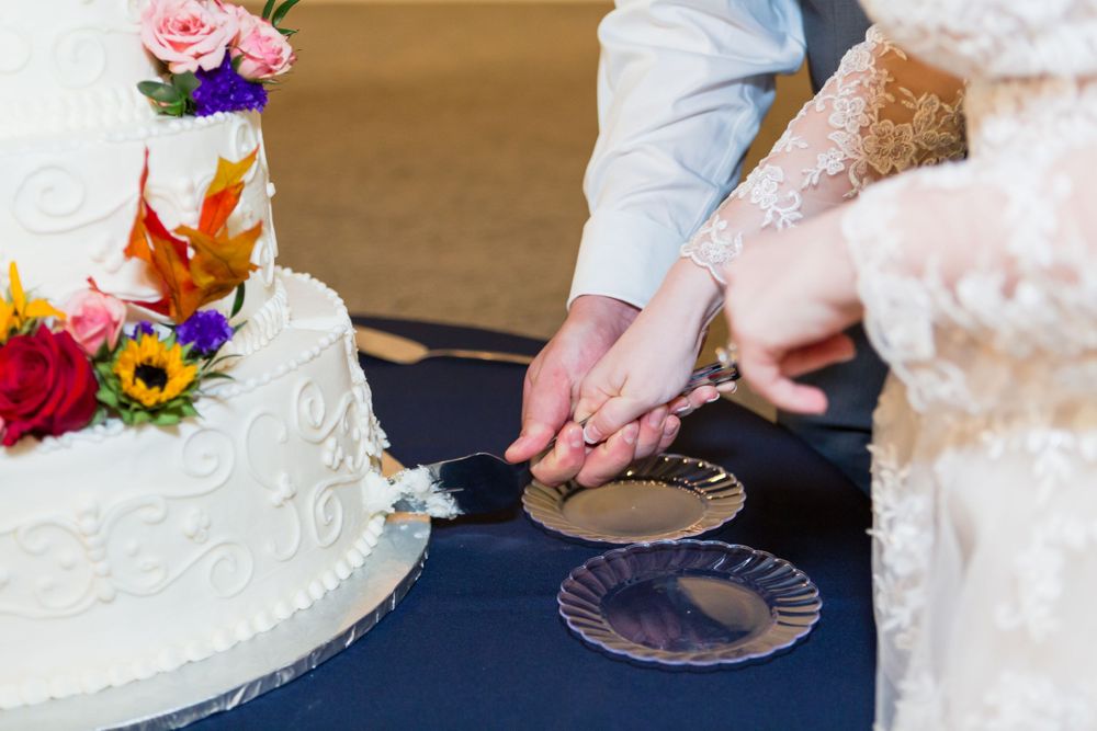 White wedding cake with colorful floral topper being cut on a navy blue tablecloth.