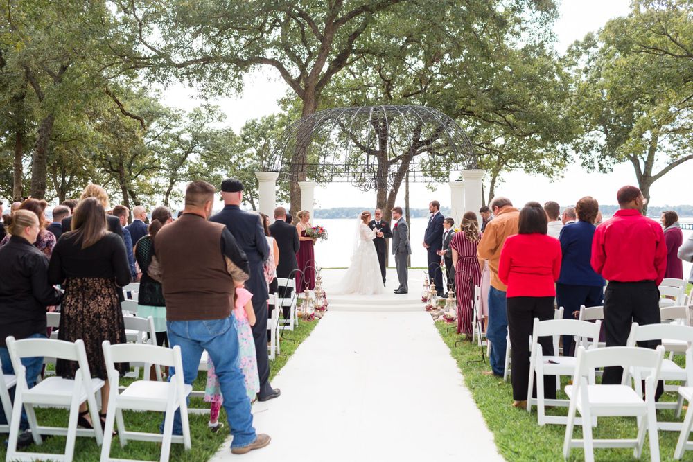 Wedding guests seated in white chairs along outdoor ceremony aisle with water view.