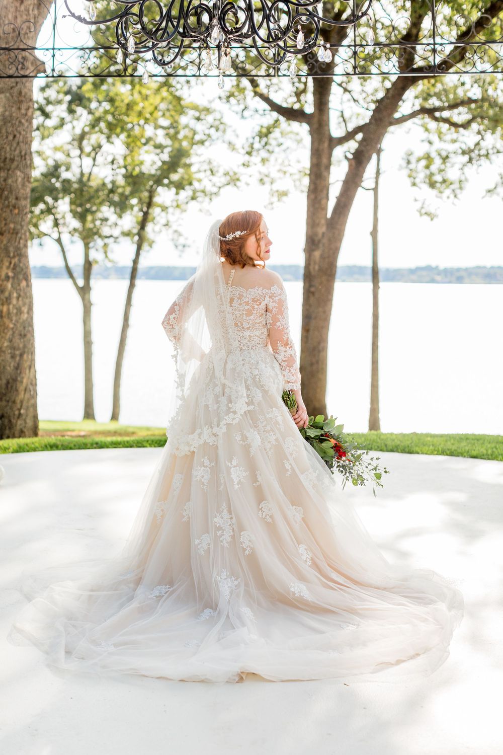 Elegant lace wedding dress with long train photographed from behind against lakeside backdrop.