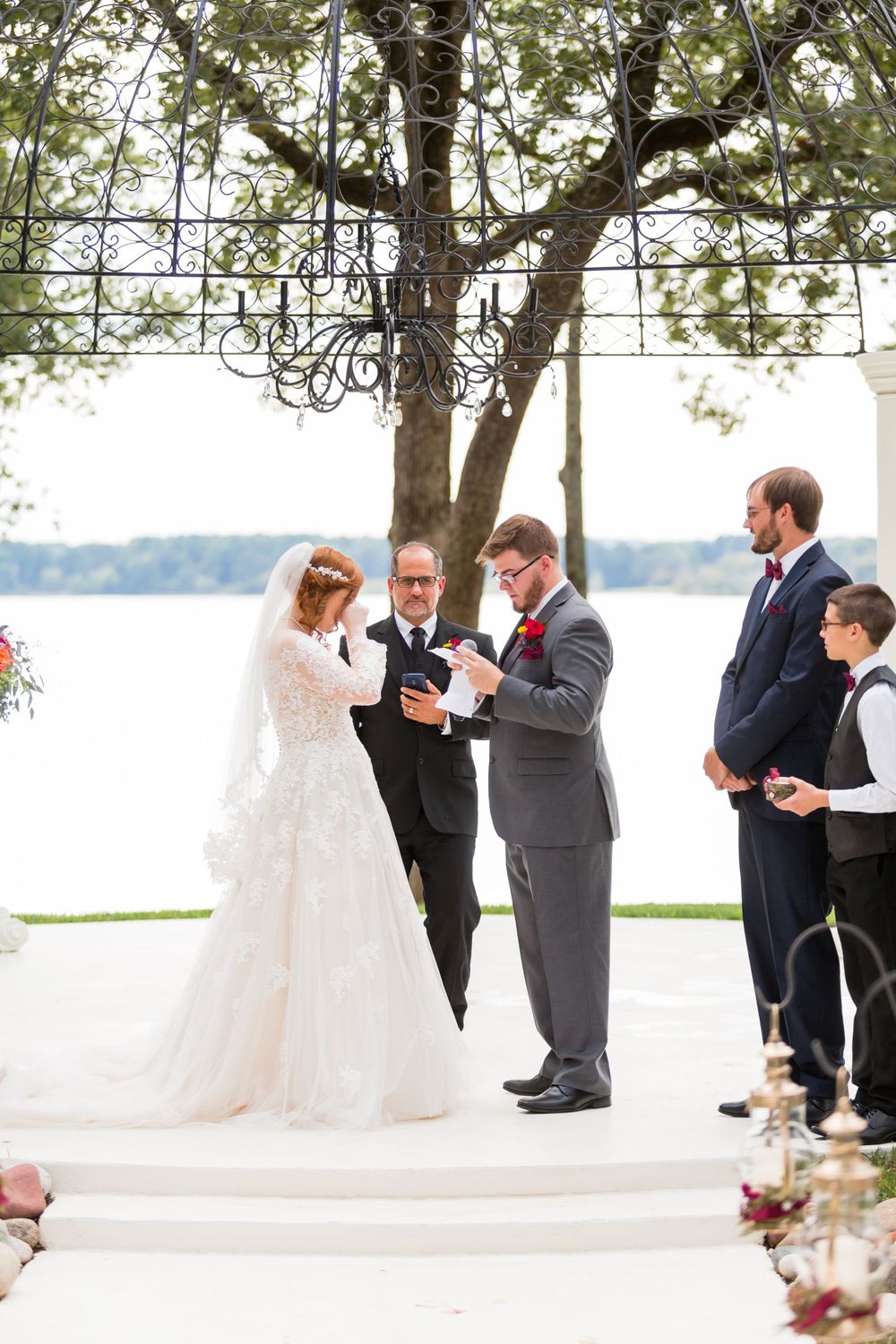 Outdoor lakeside wedding ceremony under an ornate metal pergola with a stunning waterfront backdrop.