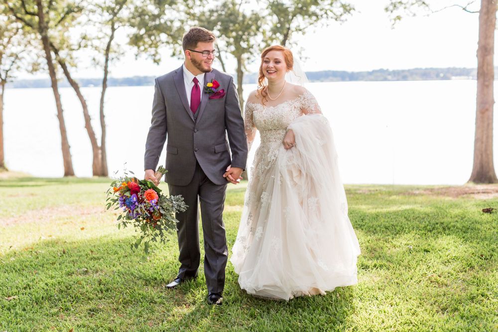 A couple walks together on a grassy lawn by a lake, with the bride holding a vibrant purple and blue bouquet.