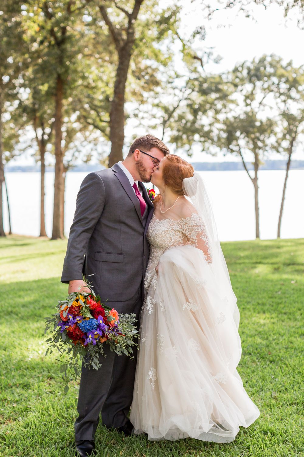 Newlyweds share an intimate moment outdoors with a lake view and trees in the background.