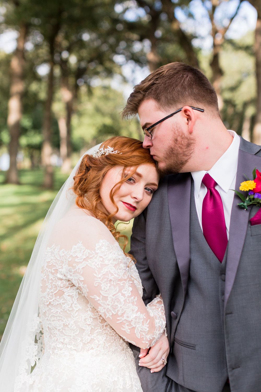 A tender wedding moment captured between the couple outdoors with sunlit trees in the background.