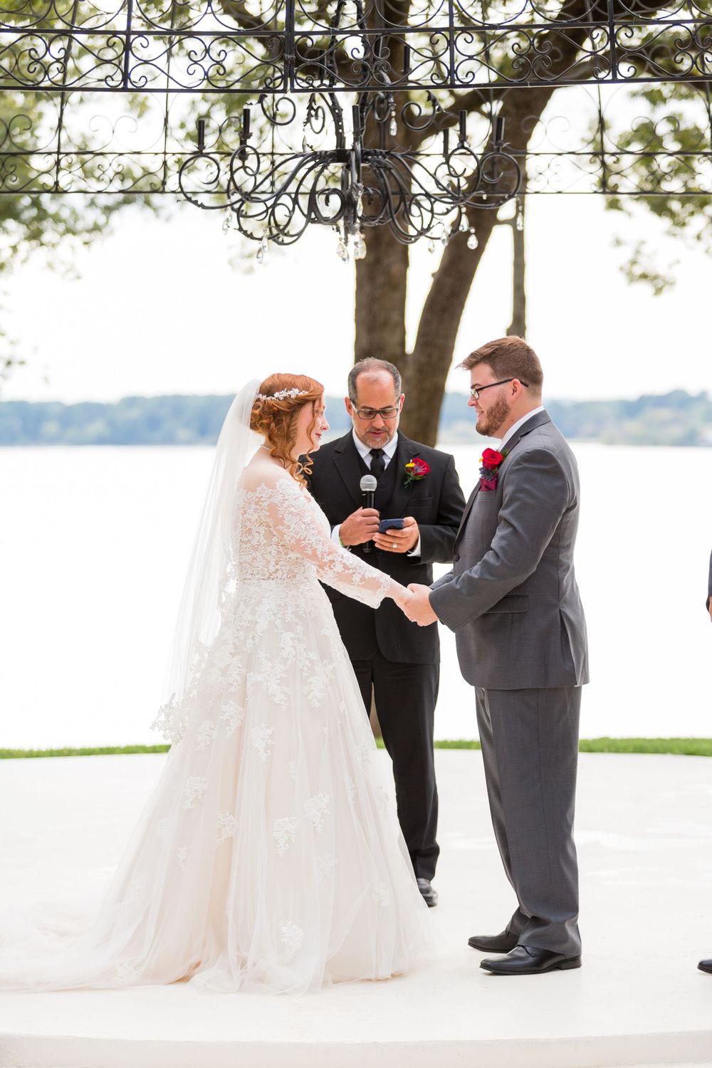 Wedding ceremony taking place on a white platform by a lake with an elegant metal arbor overhead.