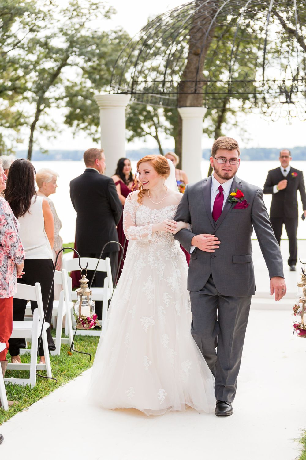 Happy newlyweds walking down white aisle after outdoor lakeside ceremony.