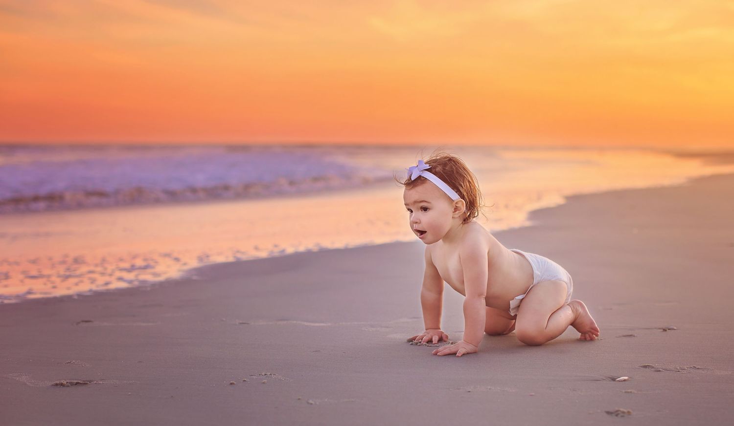 Baby crawling on beach at sunset with orange sky and ocean waves in background.
