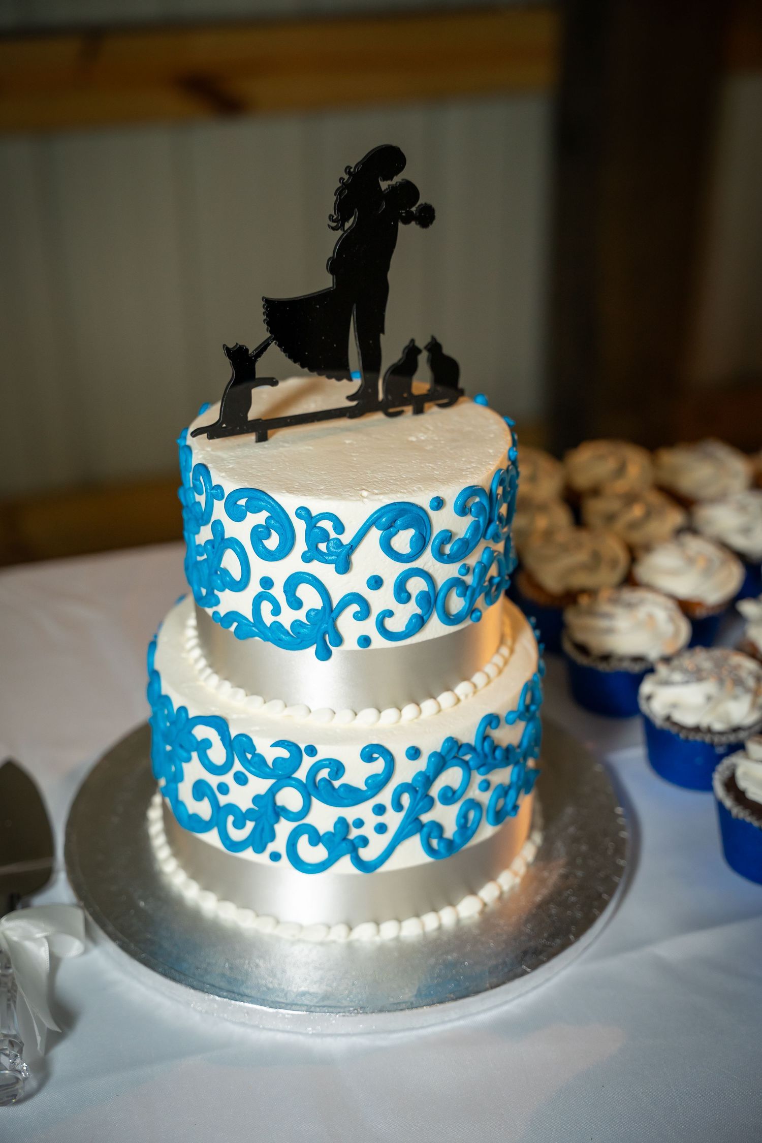 Two-tier white wedding cake with blue scroll designs and silhouette cake topper featuring a couple and pets.