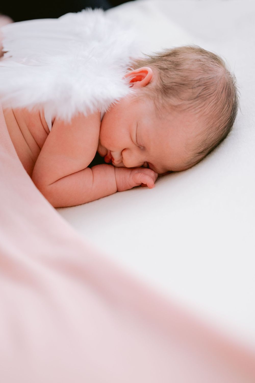 Sleeping baby covered with white fluffy blanket on light pink backdrop.