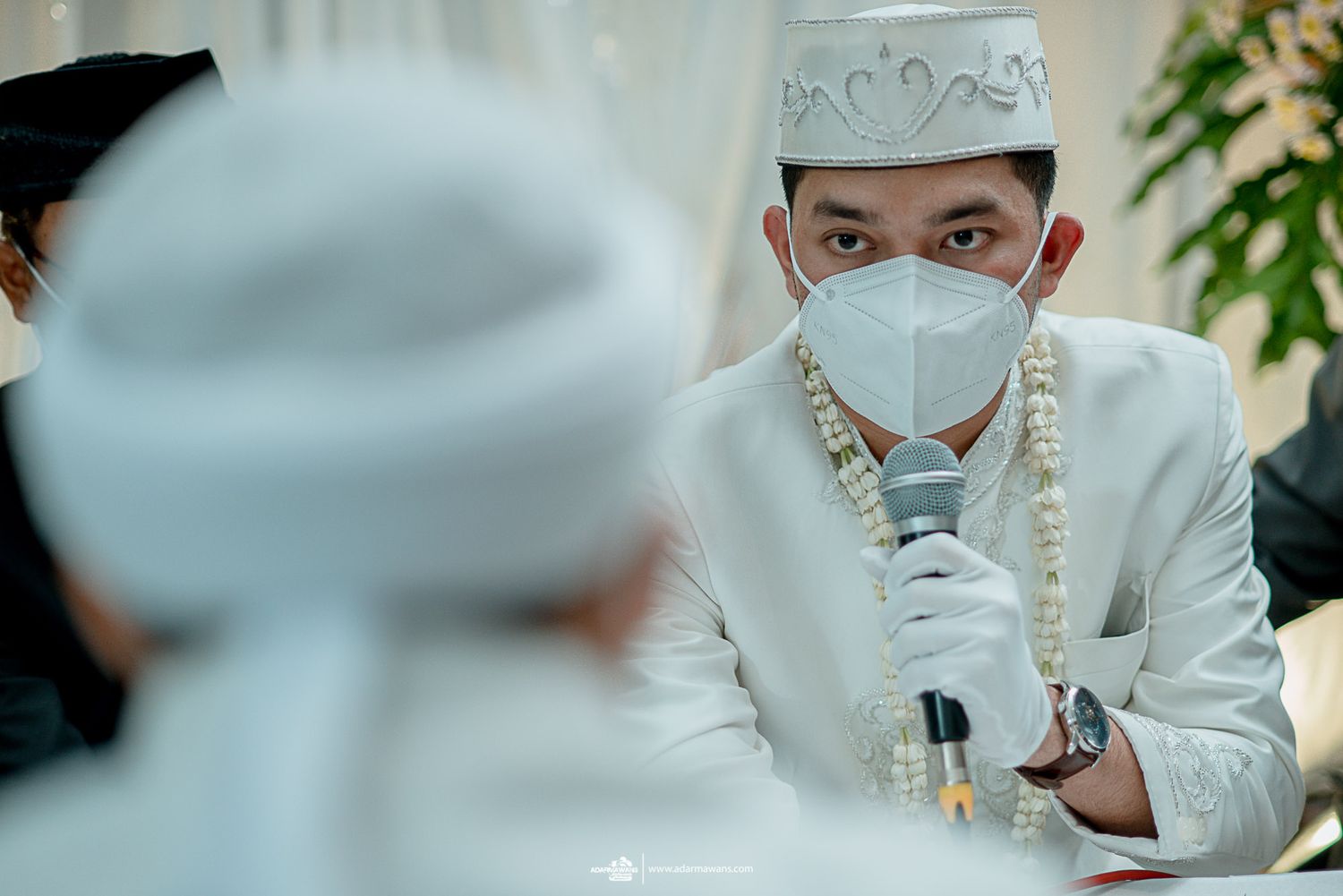 Person in white attire and face mask speaking into microphone during traditional ceremony.