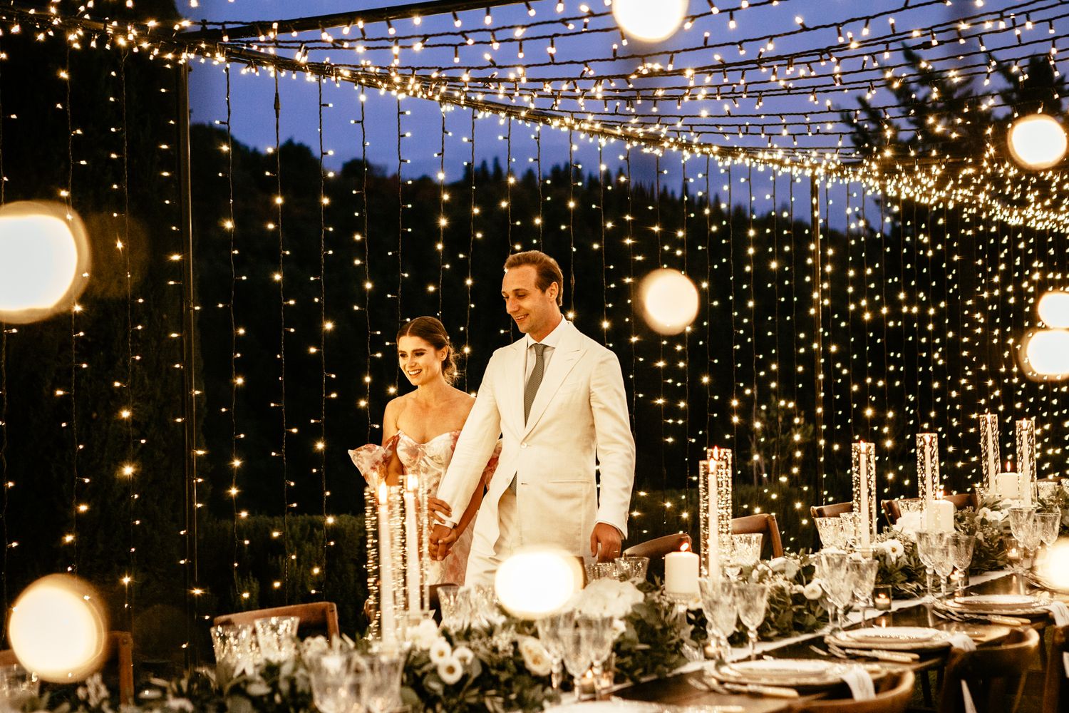 couple walk under a canopy of fairy lights next to the table set for their wedding breakfast