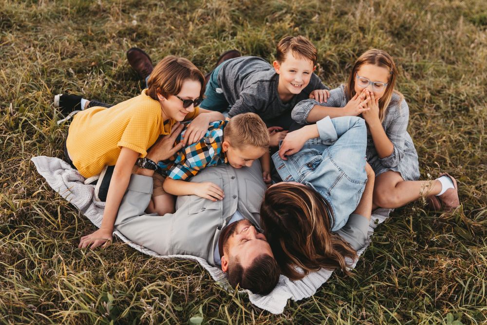 A family shares in laughter as they lie together on a blanket in the grass. Mom and Dad are on bottom with kids piled on