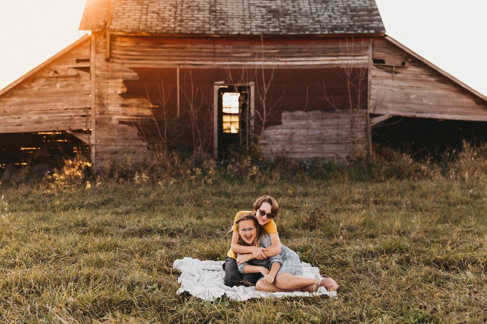 Two sisters hug one another on a blanket in front of a rustic barn structure. Golden light glows in the background.