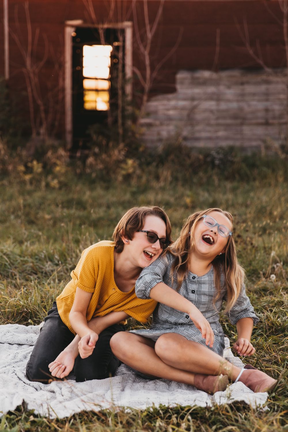 Two sisters share in laughter as they sit side-by-side on a blanket. They're both white and one wears glasses.