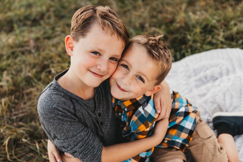 Two brothers smile at the camera as they share an embrace. Their cheeks are pressed against one another.