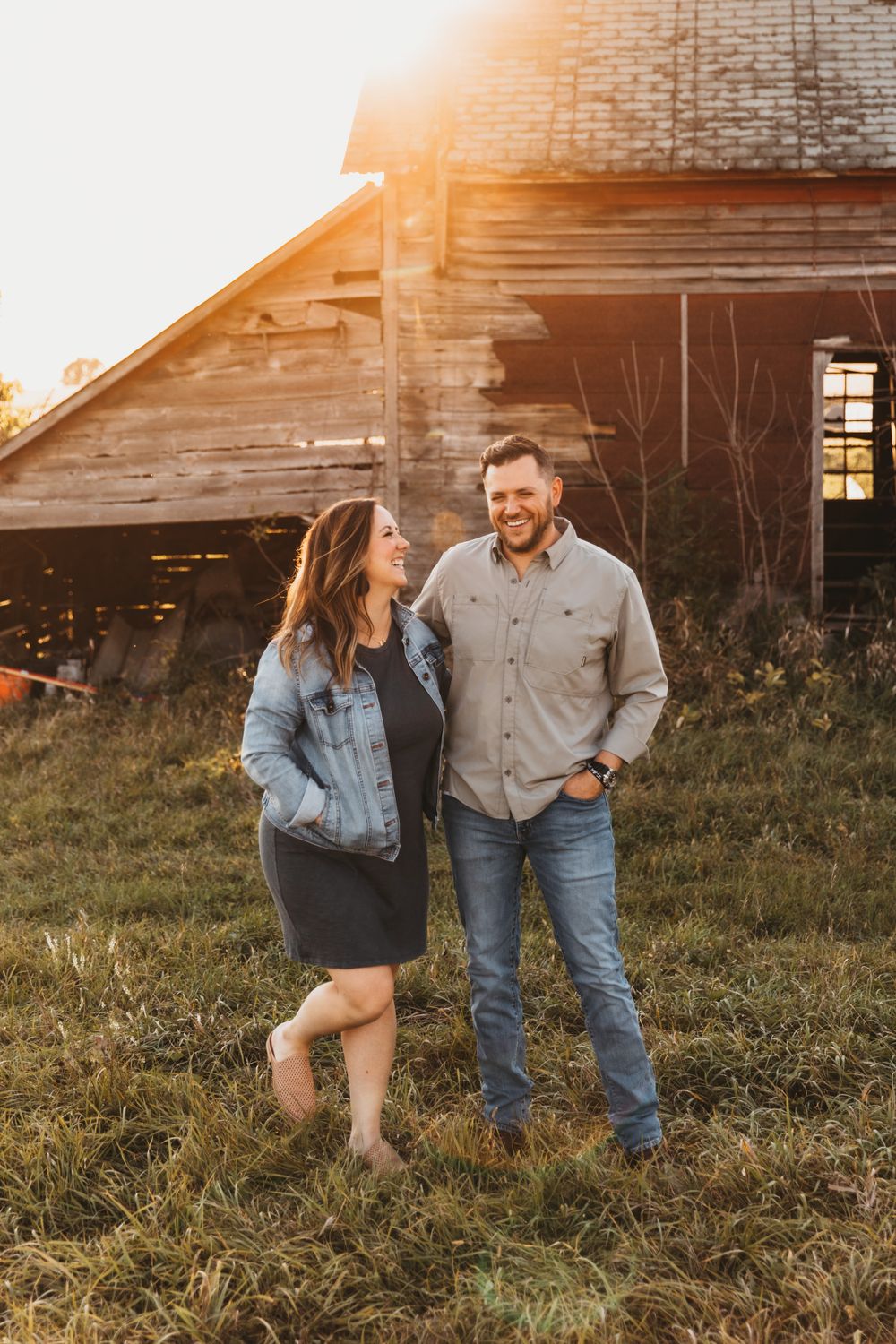 A husband and wife share in laughter as they stand in front of a rustic barn structure. Golden light glows behind them.