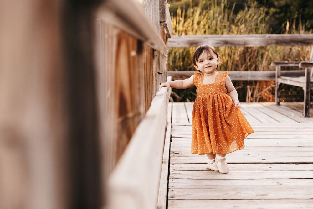 A one-year old girl stands on a wooden dock supported by her hand holding onto a wood beam. She wears a yellow dress.