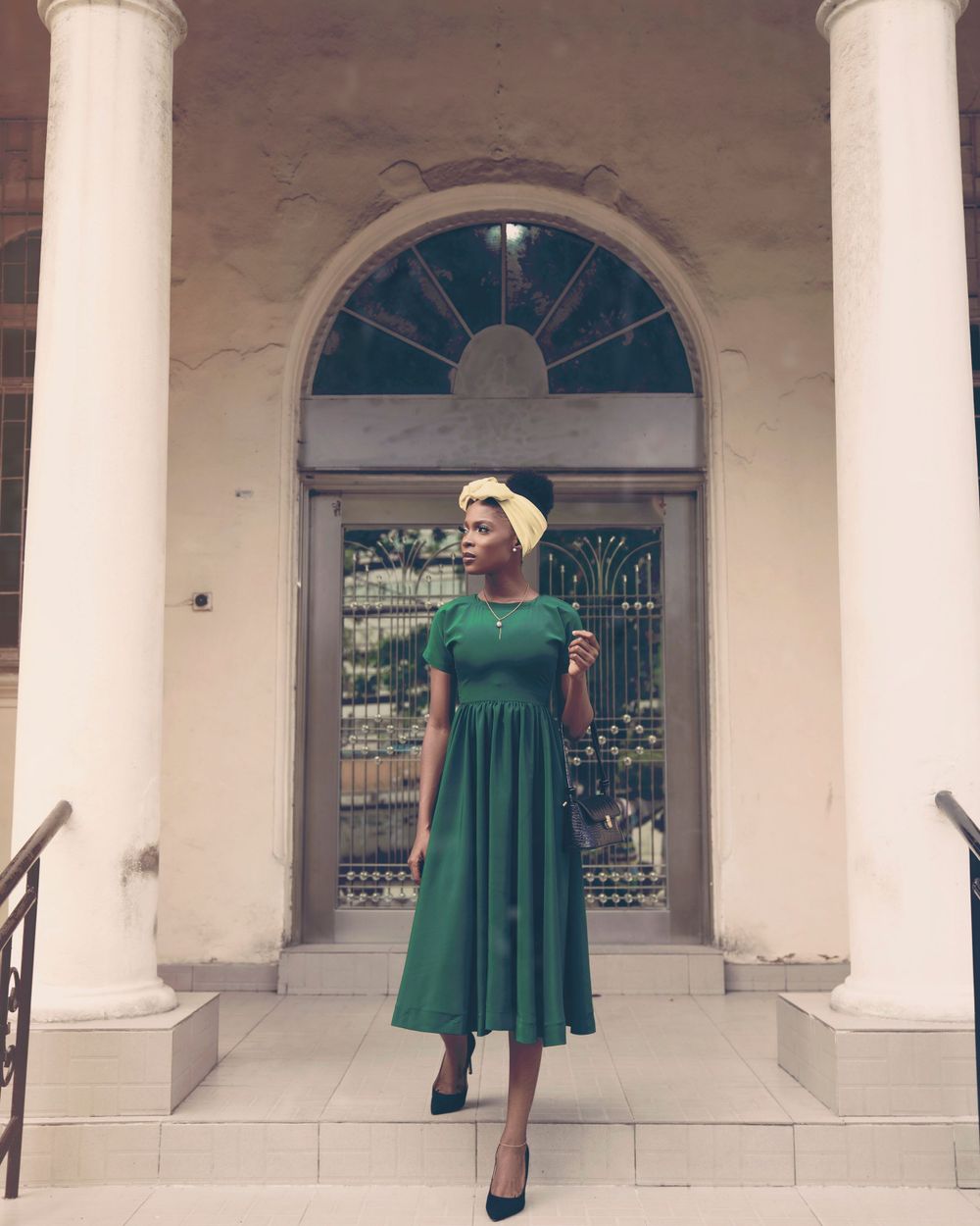 A woman in a green midi dress and yellow headband poses elegantly on steps beneath a classical arched entrance.