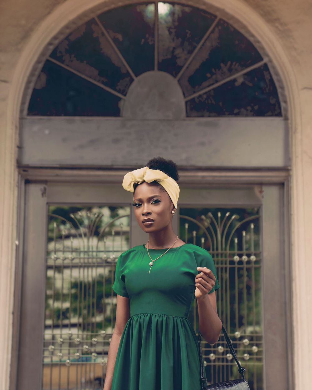 A vintage-inspired portrait of a woman in an emerald green dress and yellow headband standing by an arched doorway.