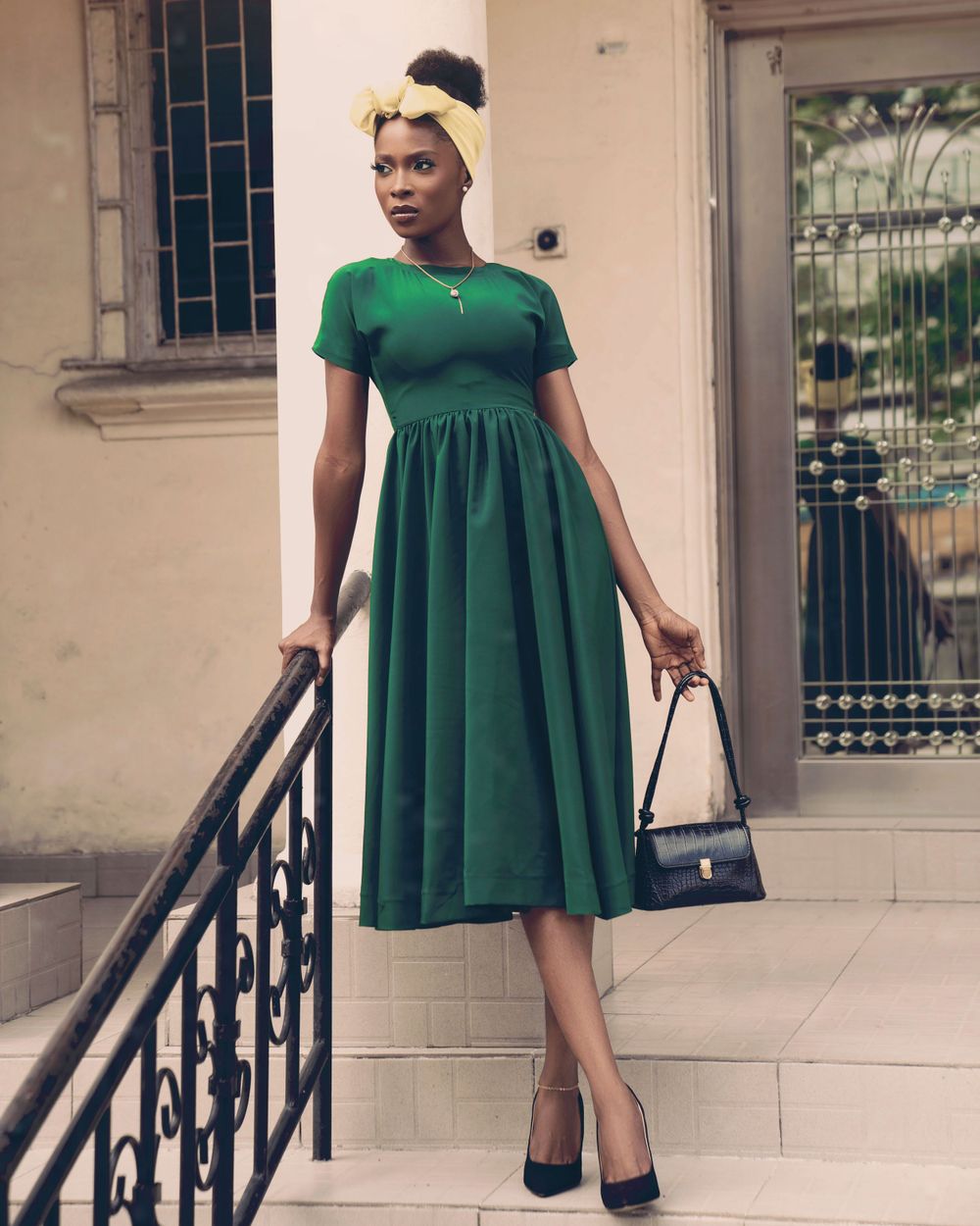 A woman in a vintage-style green dress with short sleeves stands on steps holding a black handbag.