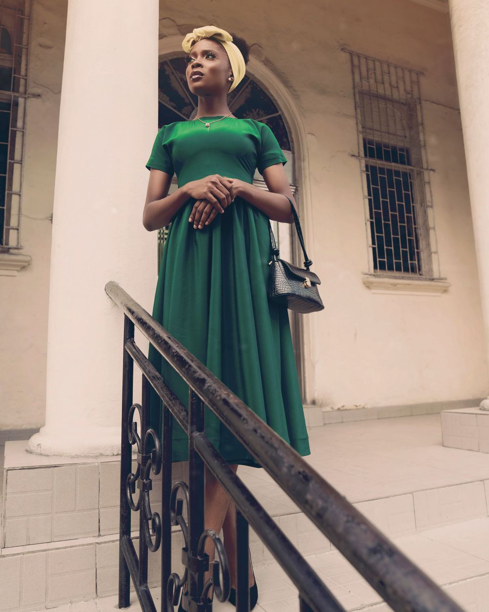 A stylish woman in a green tea-length dress poses on stone steps with ornate iron railings.