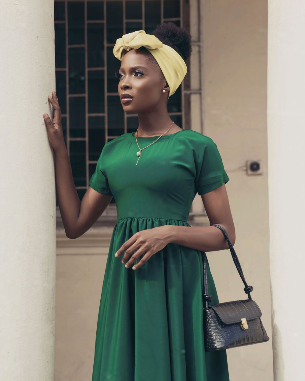 A woman in an emerald green dress and yellow head wrap poses against a textured wall.