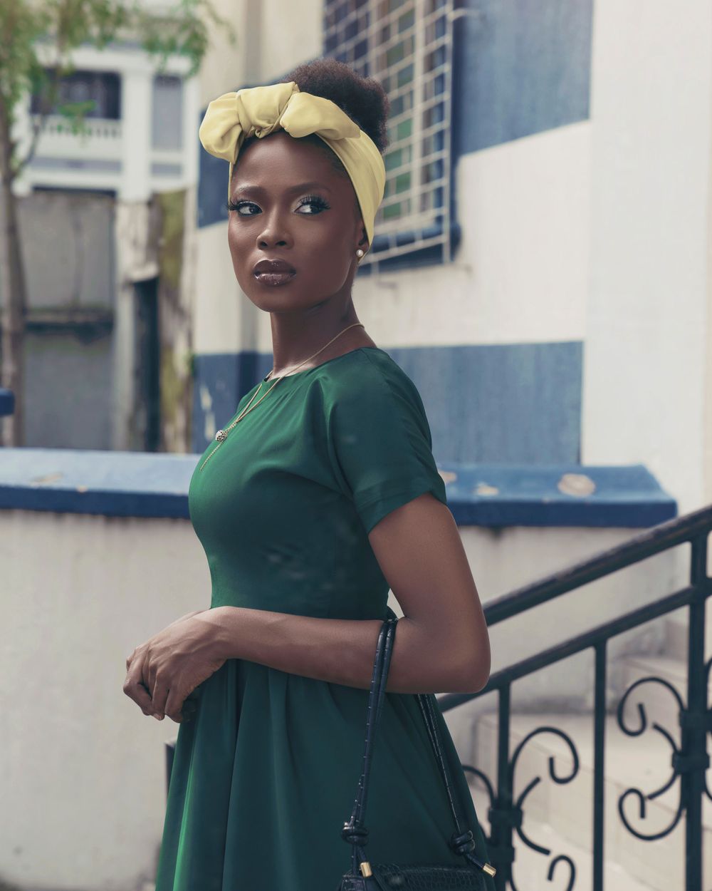 A woman in a green dress and yellow head wrap poses against a striped blue and white wall.