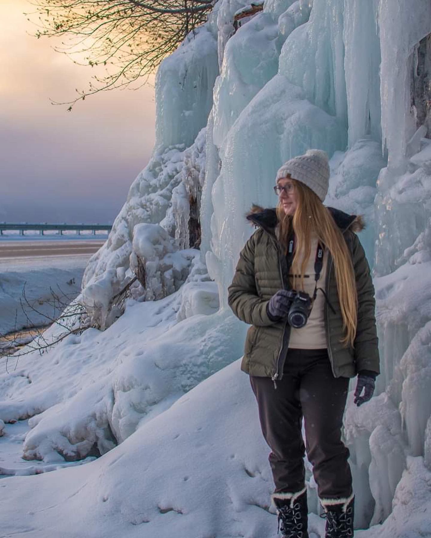 Person in winter gear stands by dramatic ice formations at sunset along frozen shoreline.