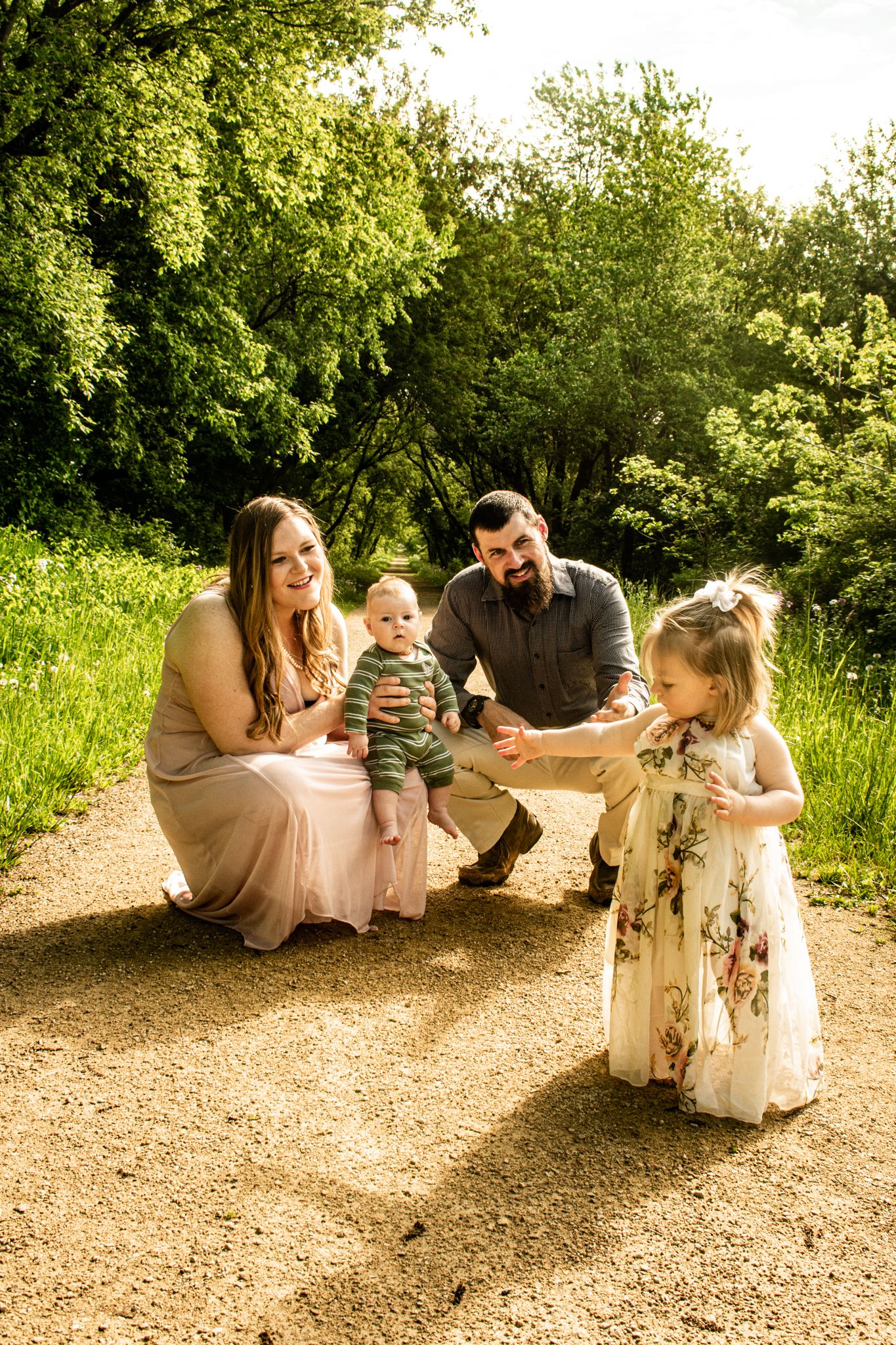 A sunny family portrait on a dirt path surrounded by lush green summer foliage.