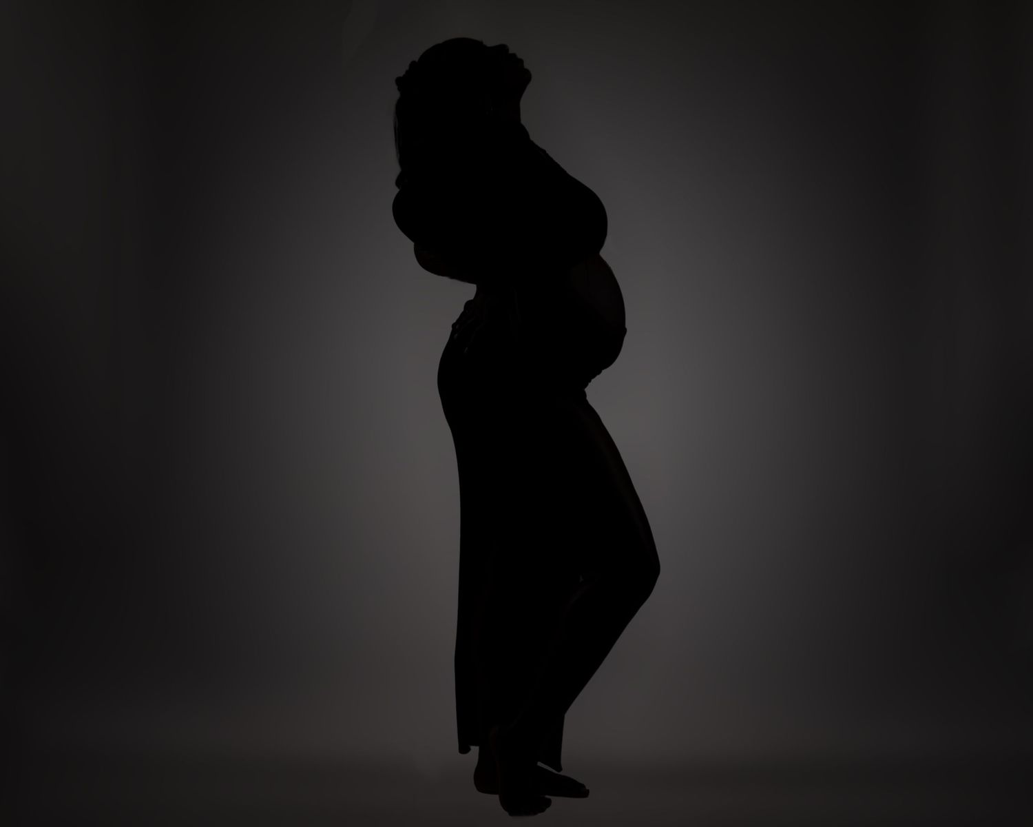 A silhouetted side view of a pregnant woman standing against a dark background.