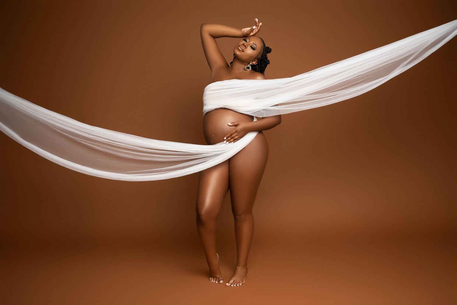 Artistic studio maternity portrait with flowing white fabric draping against warm brown background.