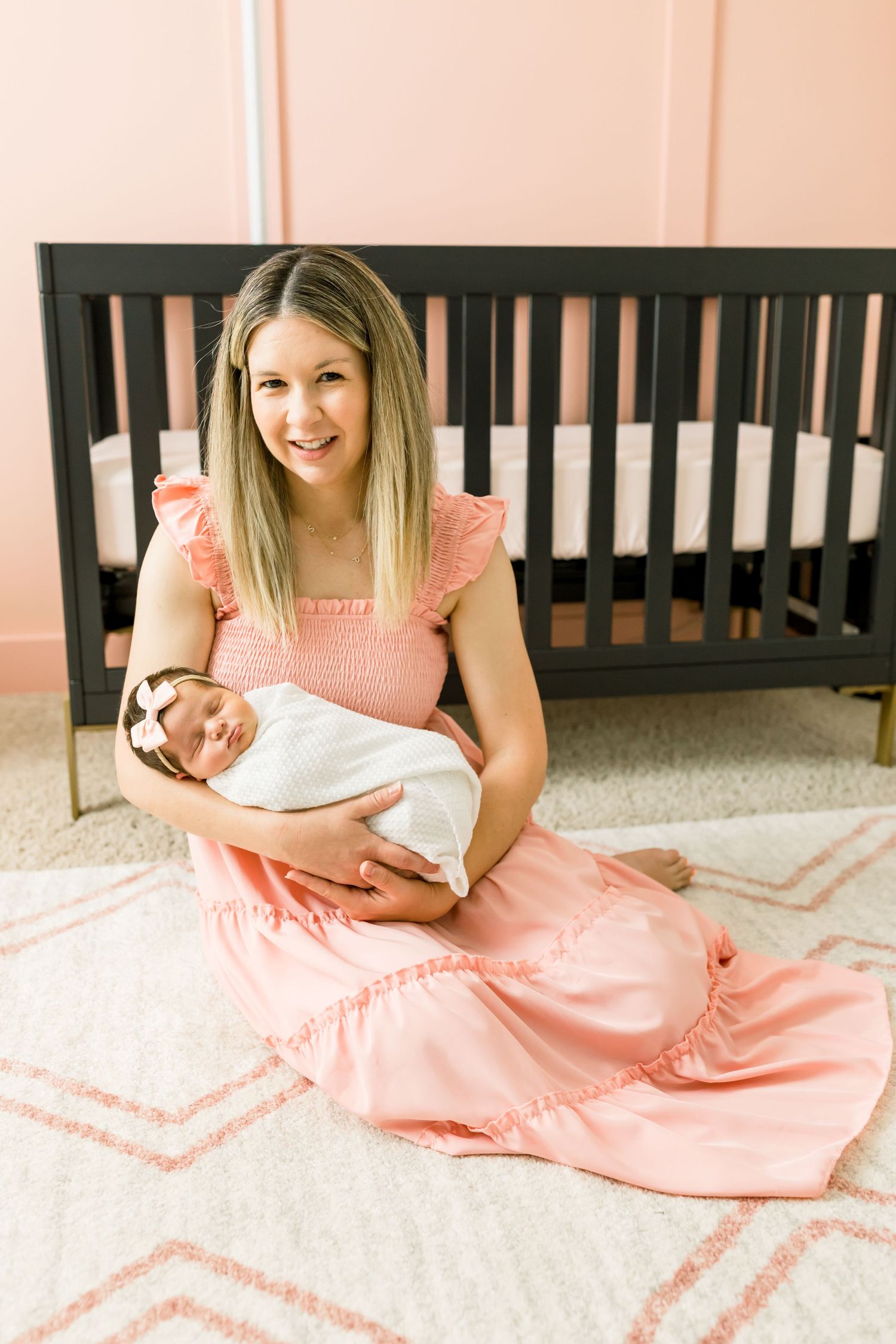 Nursery setting with dark wood crib and person in pink dress holding swaddled infant.