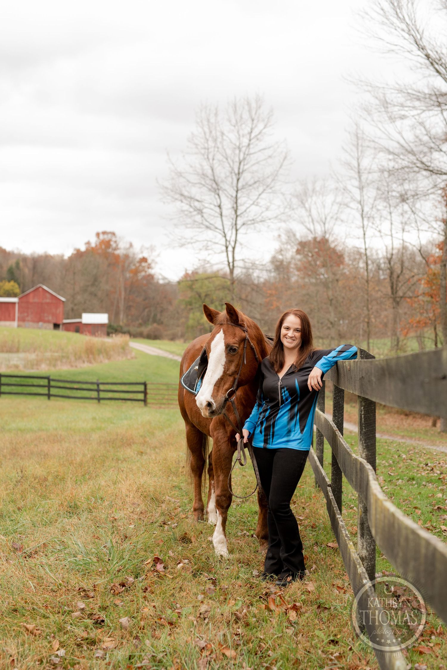 A chestnut horse with white markings stands by a fence with autumn trees and red barn in background.