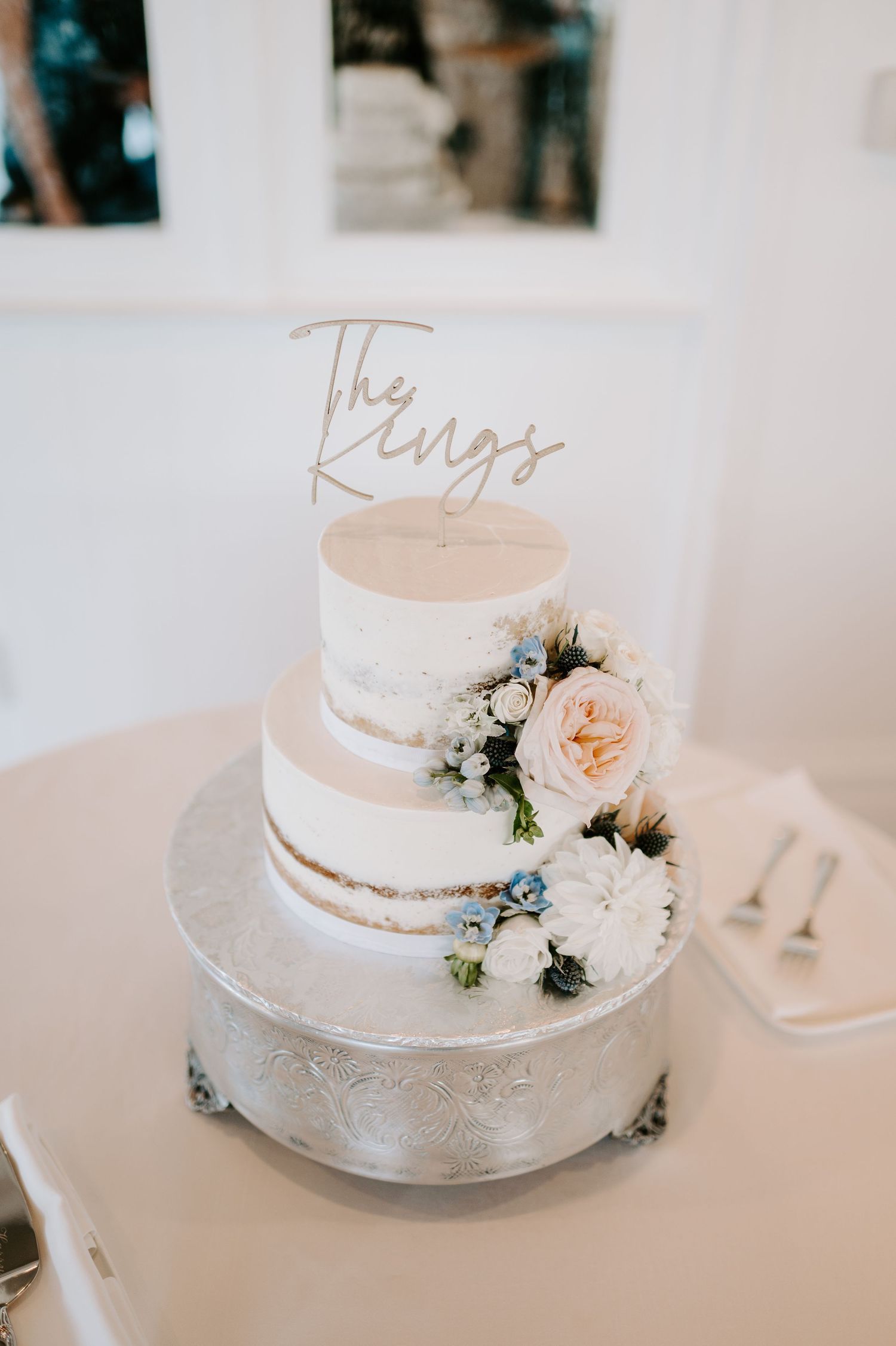 A simple white wedding cake decorated with delicate flowers and metallic text on top.