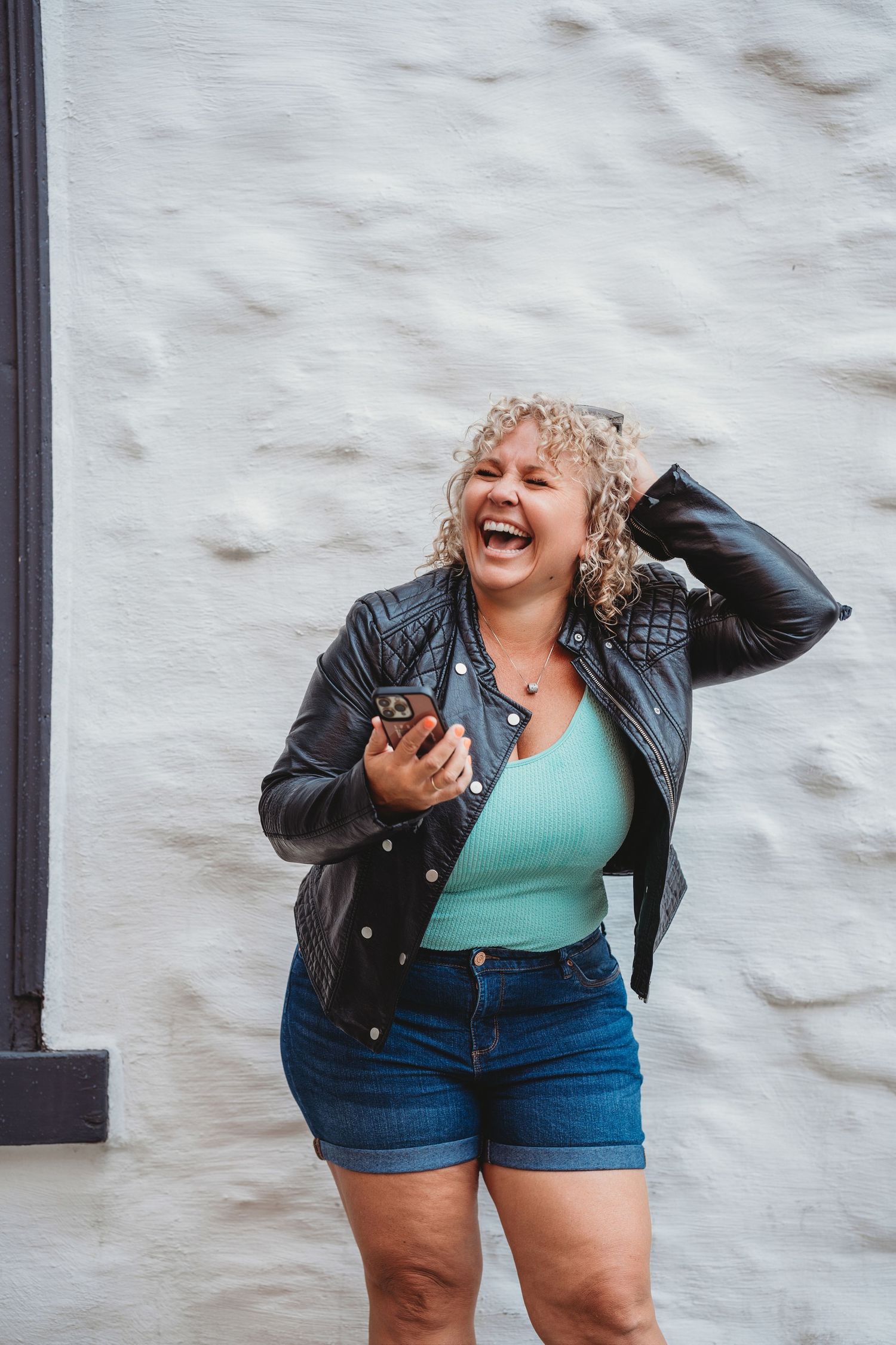 Woman in turquoise top and leather jacket laughing joyfully against white wall.