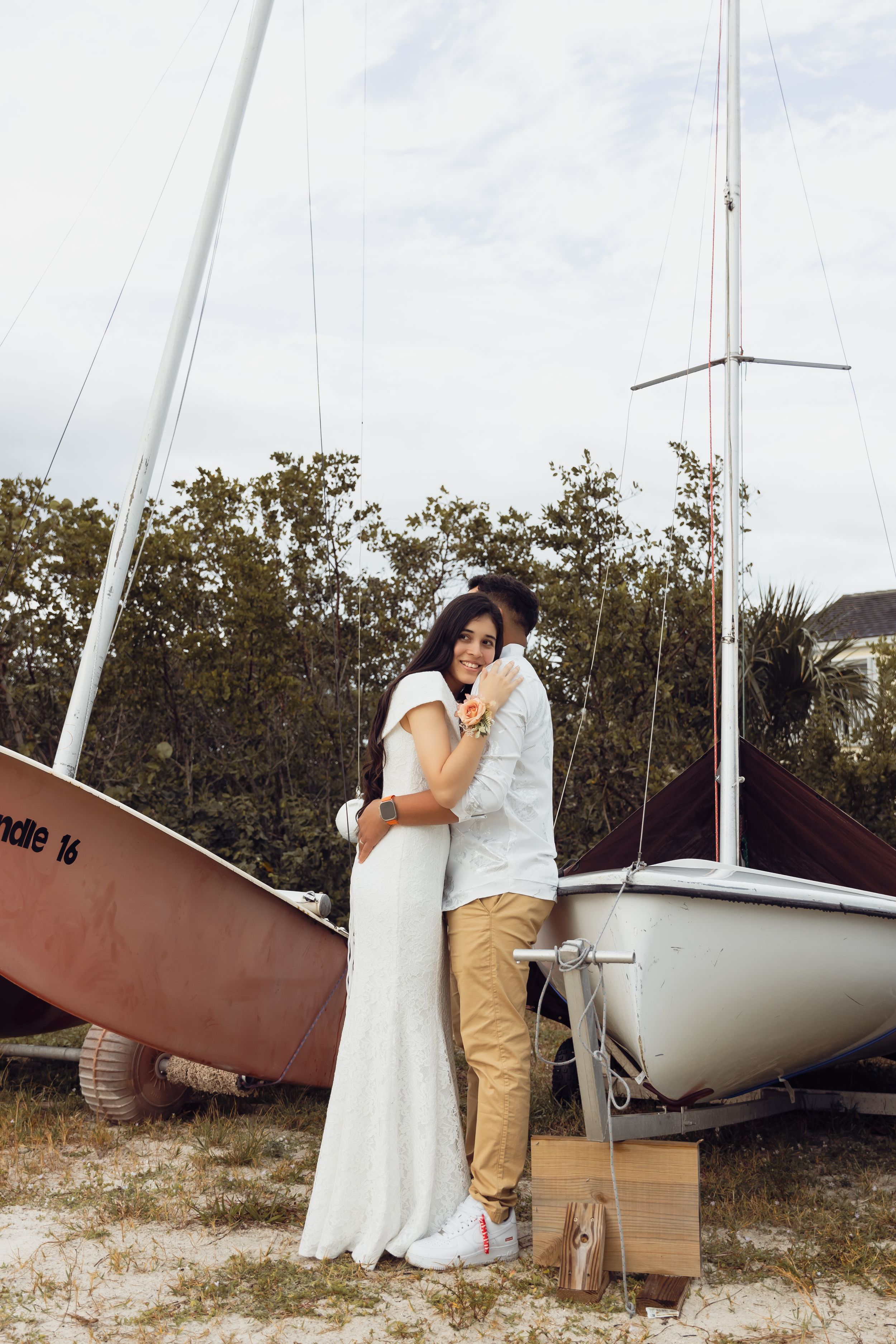 Couple in formal attire posing together near sailboats on shore.