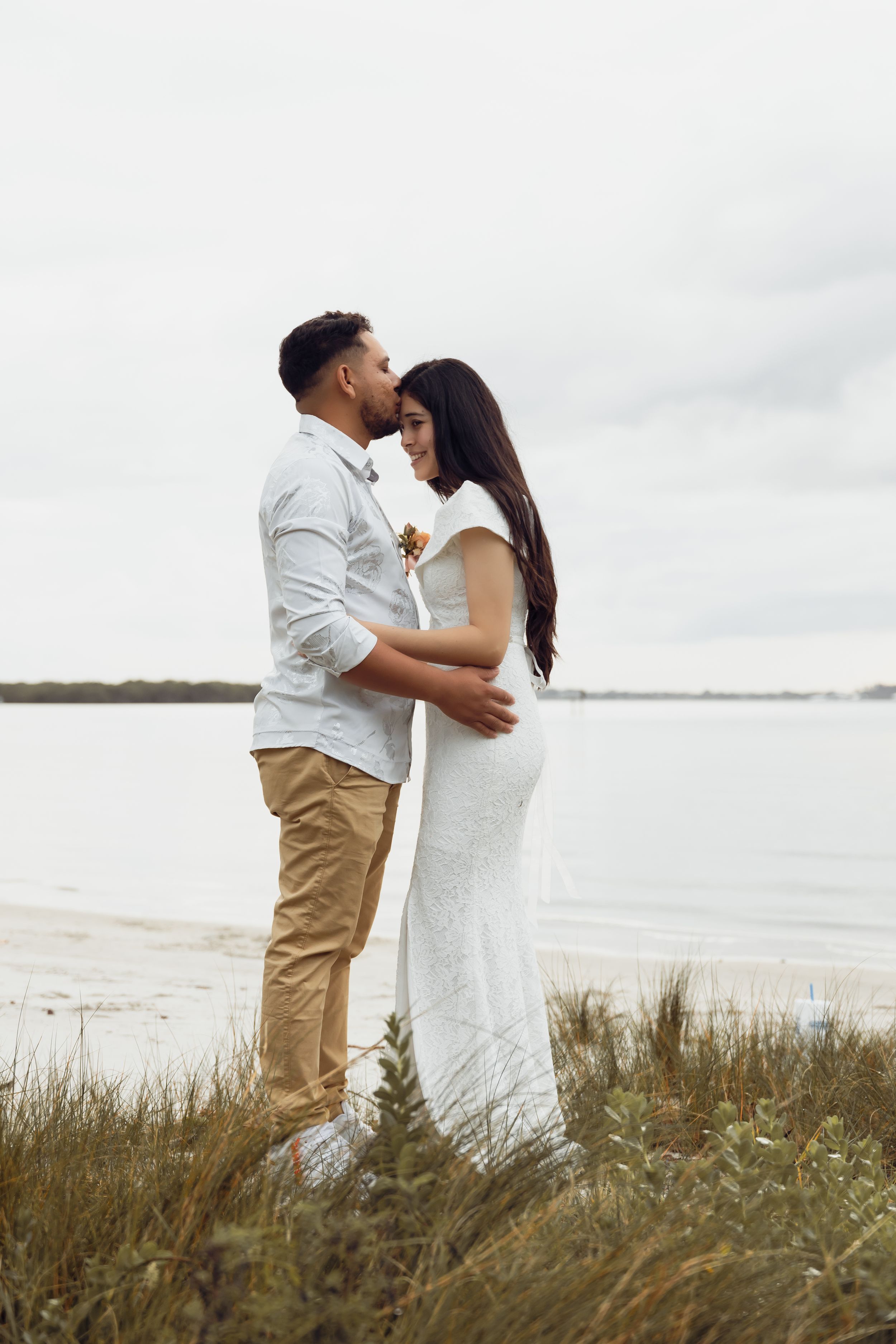 Couple embracing on coastal grassland wearing white and khaki outfits.
