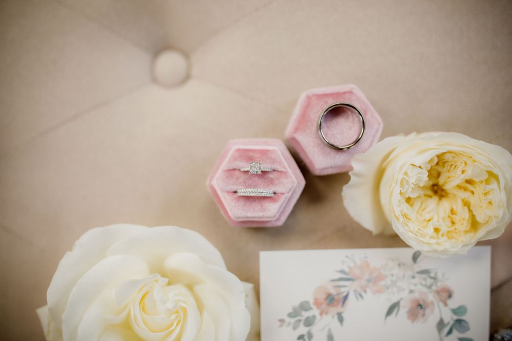 Pink hexagonal ring boxes and white roses arranged with floral stationery on beige surface.