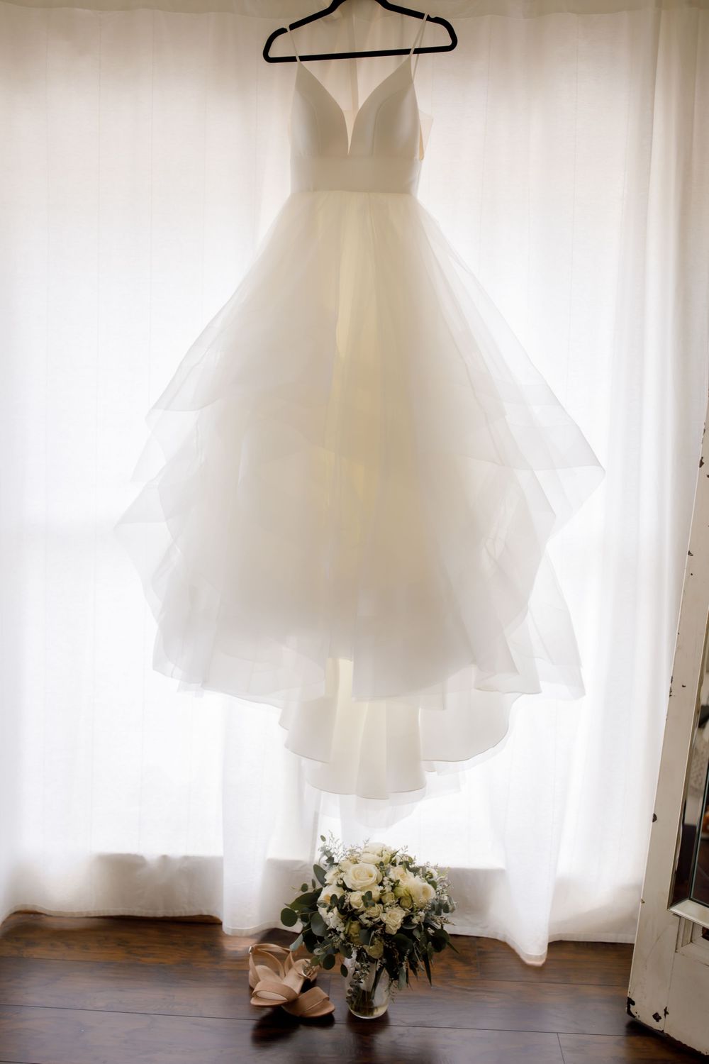 White wedding dress hanging on display with bouquet and shoes beneath against light background.