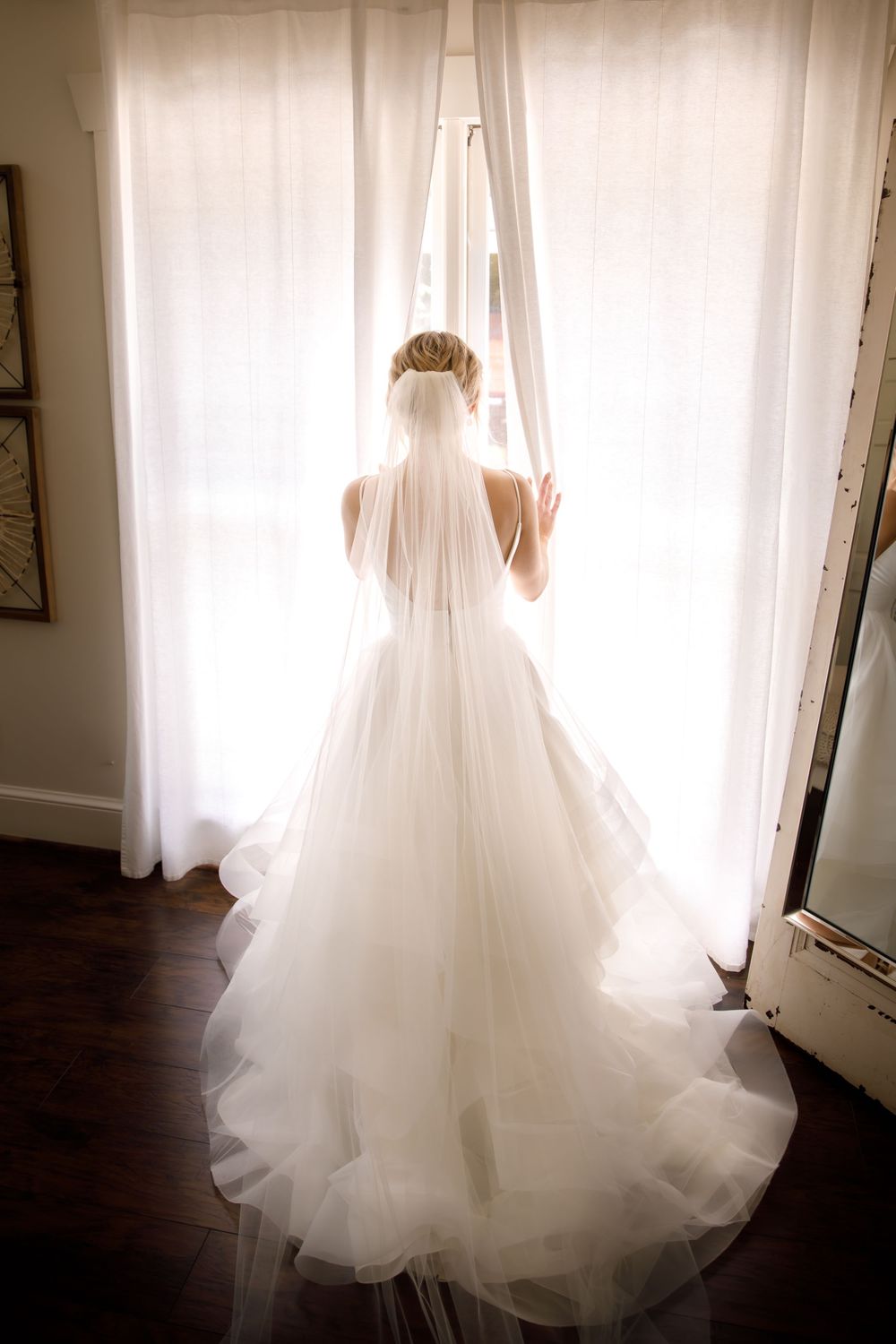 Backlit silhouette of bride in flowing wedding gown and veil standing by window.