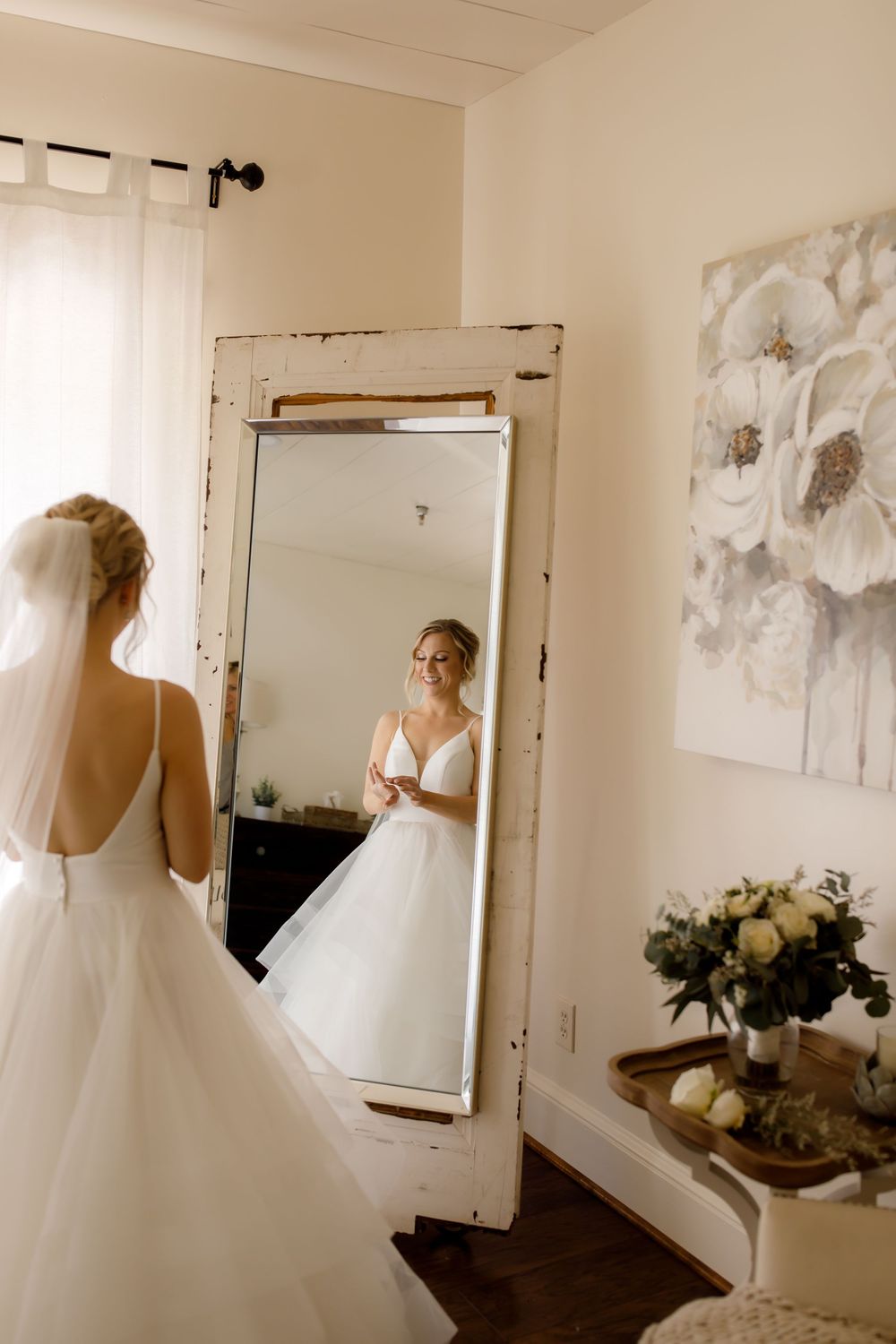 A stunning wedding dress reflection in a vintage distressed mirror with white floral artwork on the wall.