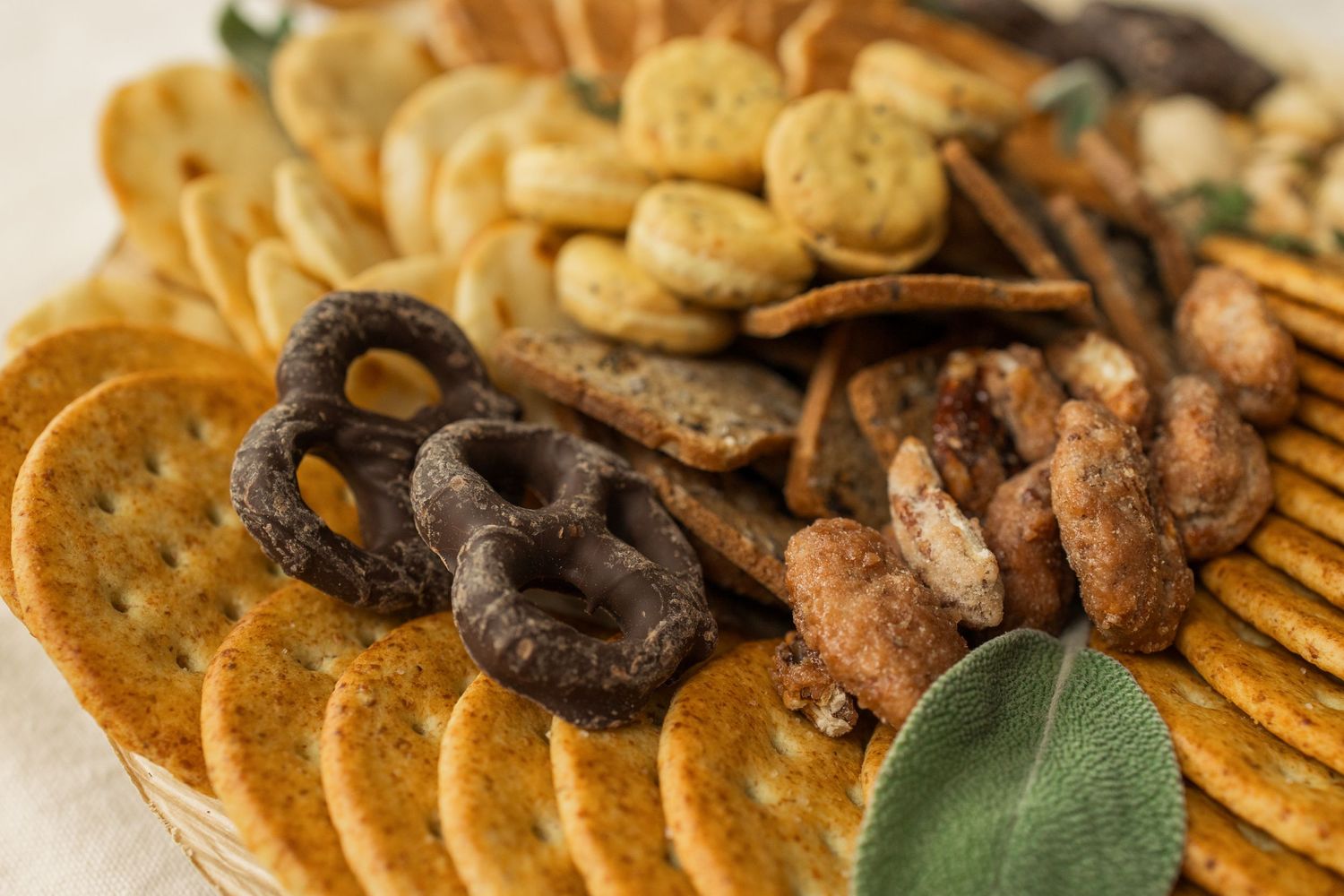 A charcuterie board with various crackers, dark chocolate pretzels and sage leaves arranged on a wooden surface.